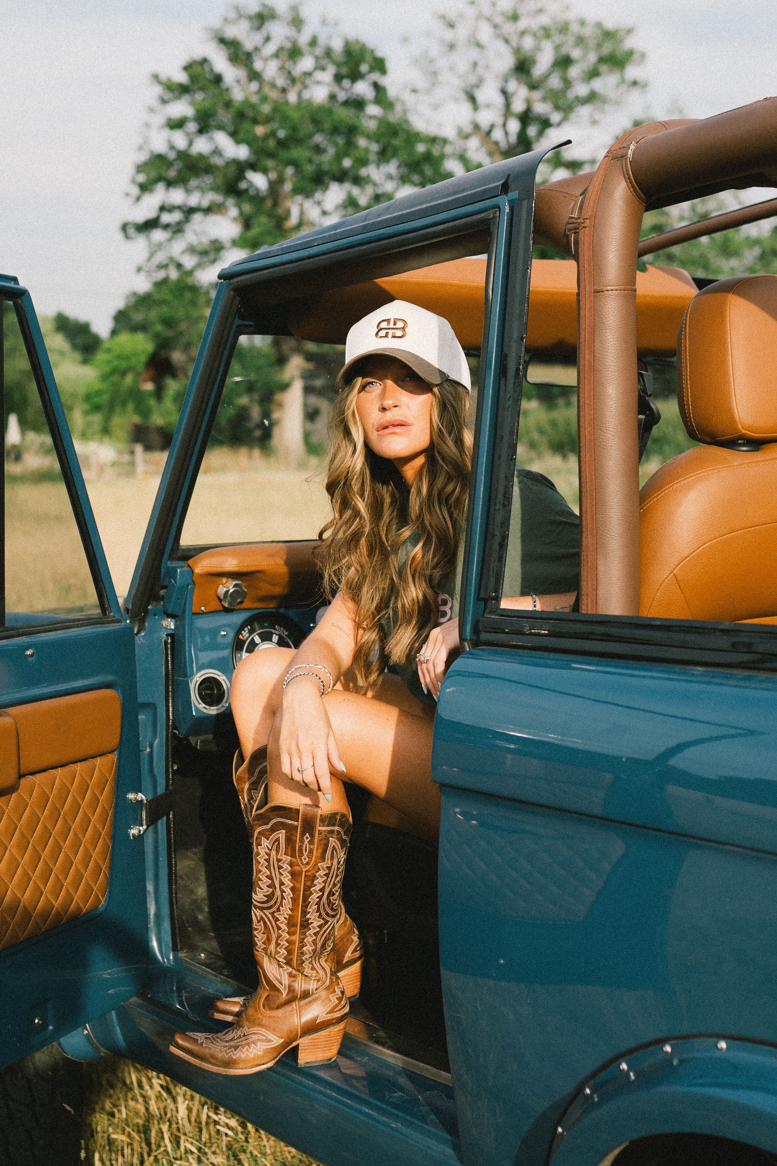 A woman wearing a white western trucker  cap with 'BB' on it and brown western boots sits in the open driver's side of a teal bronco with tan leather seats.